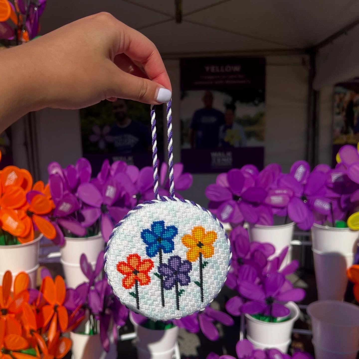Hand holding a round needlepoint ornament with floral designs against a background of potted flowers.